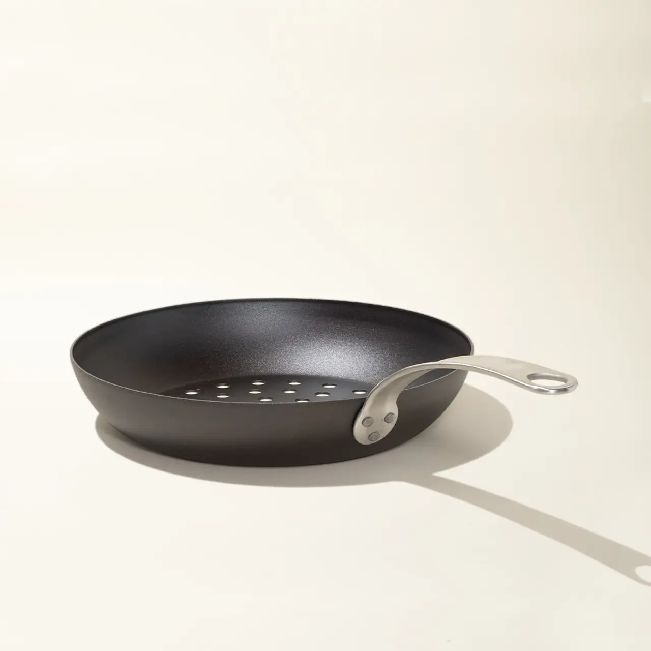 A black frying pan with a silver handle rests on a light background, casting a soft shadow to the right.