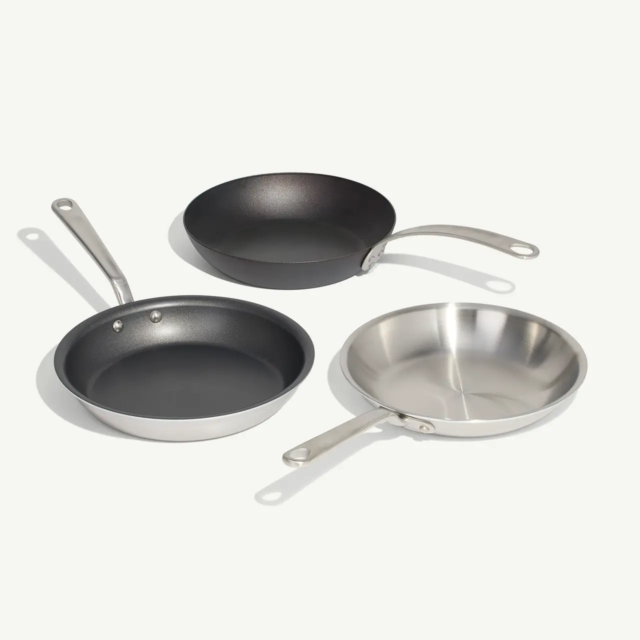 Three different types of frying pans with metal handles are arranged side by side on a plain background.