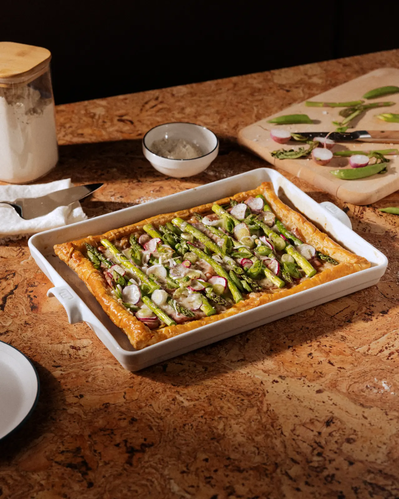 A freshly baked asparagus and cheese tart in a white dish is displayed on a countertop beside ingredients and a wooden cutting board.