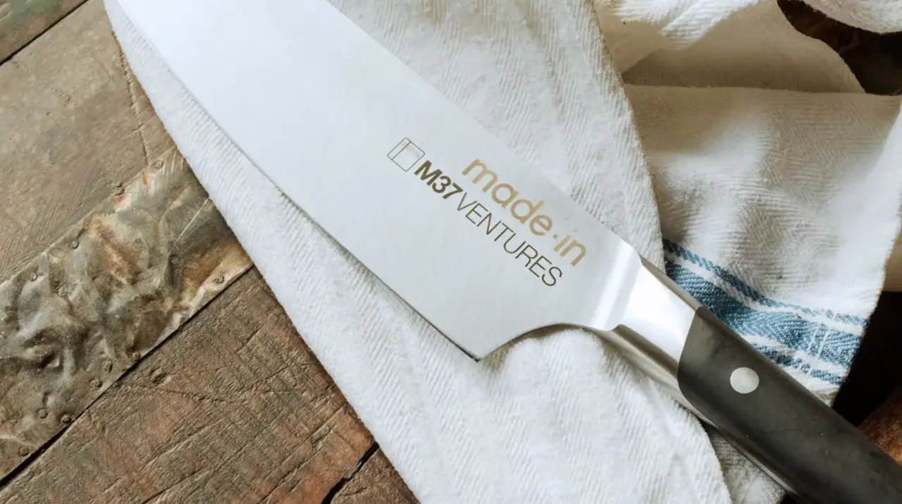 A chef's knife with the logo "made in M31Ventures" lying diagonally across a wooden surface with a striped cloth partially underneath it.