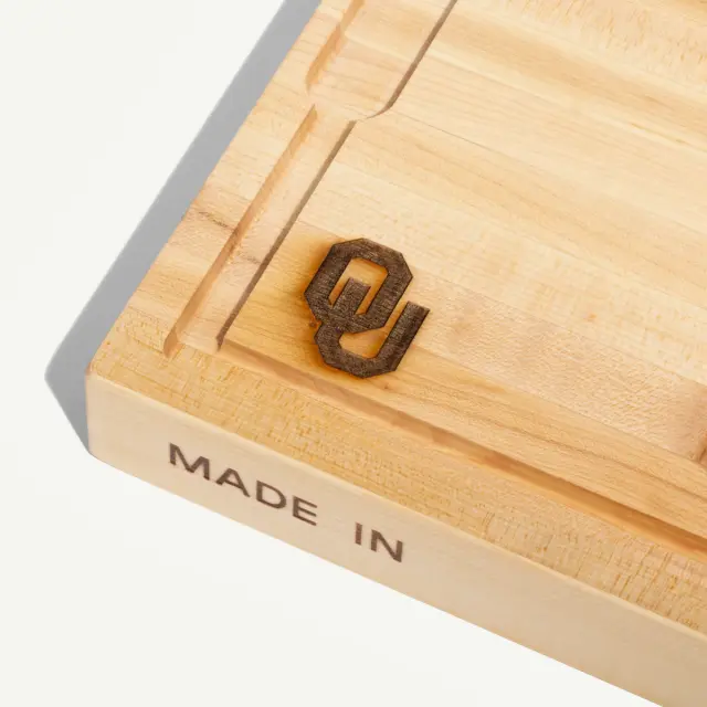 A wooden cutting board features a carved design of the letter "O" and "U," with the words "MADE IN" engraved on one side.