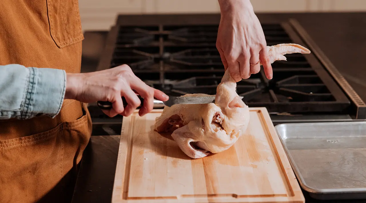 A person is preparing a chicken on a wooden board with a knife in hand.