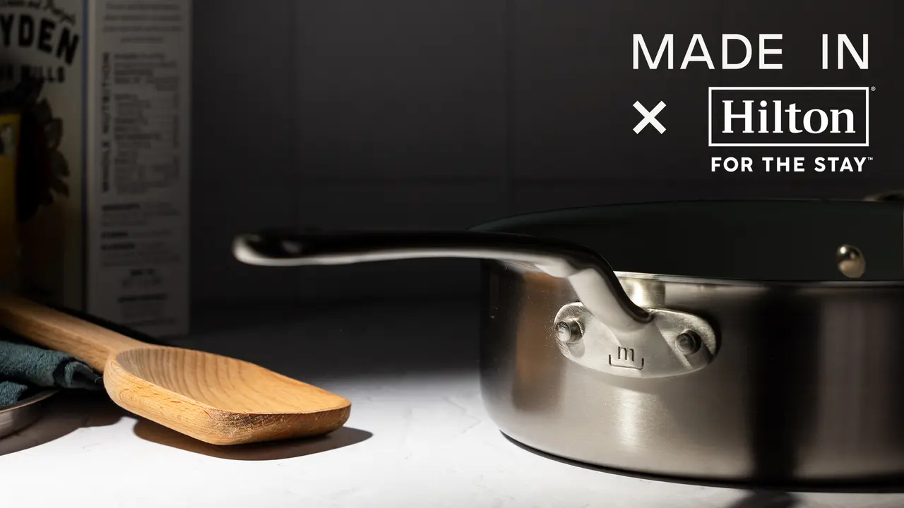 A stainless steel pan sits on a surface near a wooden spatula, with packaging reading "MADE IN X Hilton FOR THE STAY" partially visible in the background.