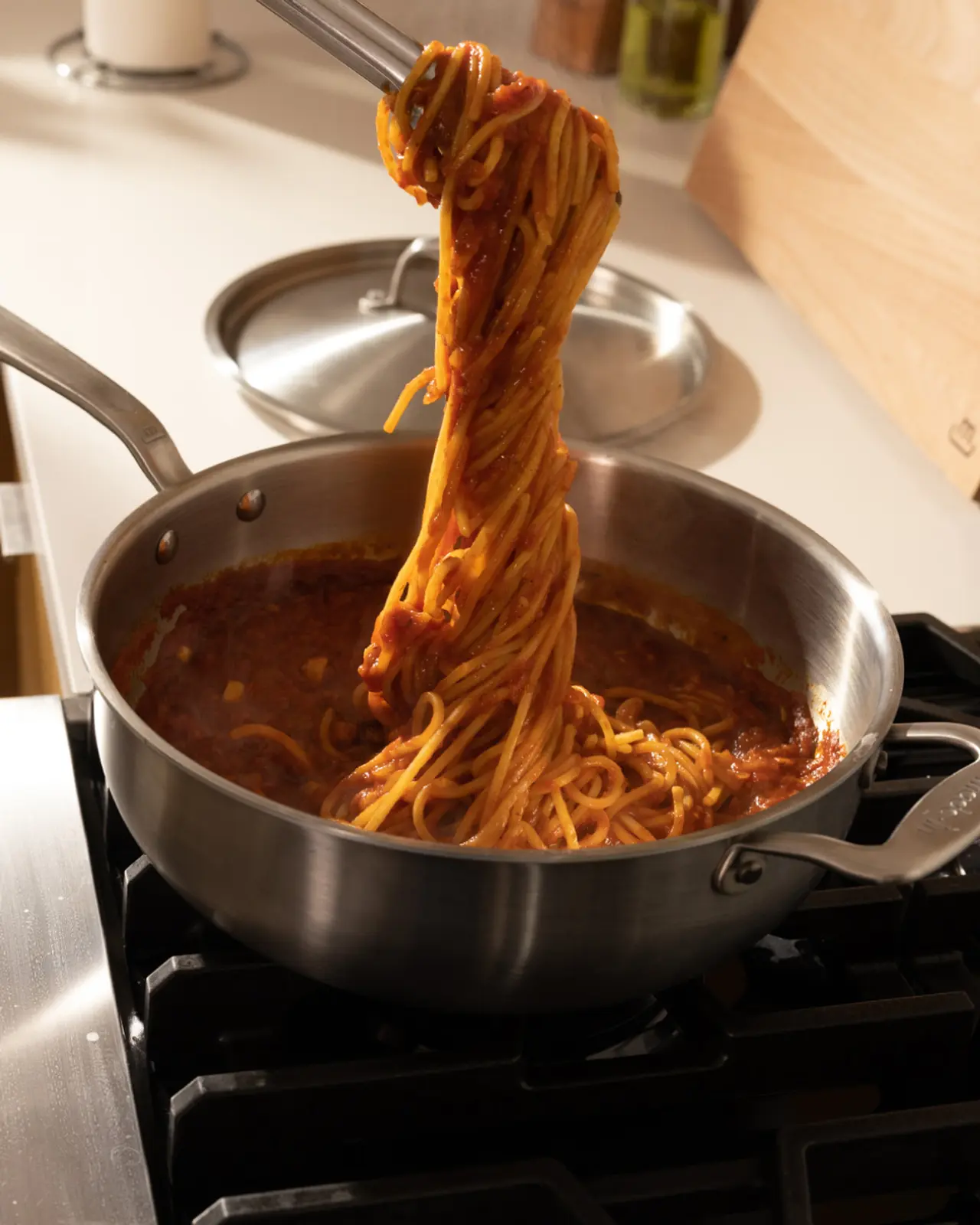 A pot on a stove containing spaghetti in tomato sauce with a large portion being lifted by tongs, showcasing the meal's thickness and richness.