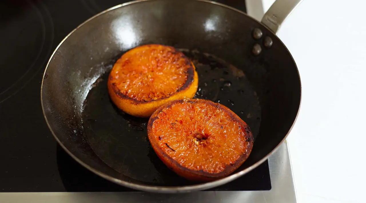 Two orange fruit halves are being caramelized in a black frying pan on a stove top.