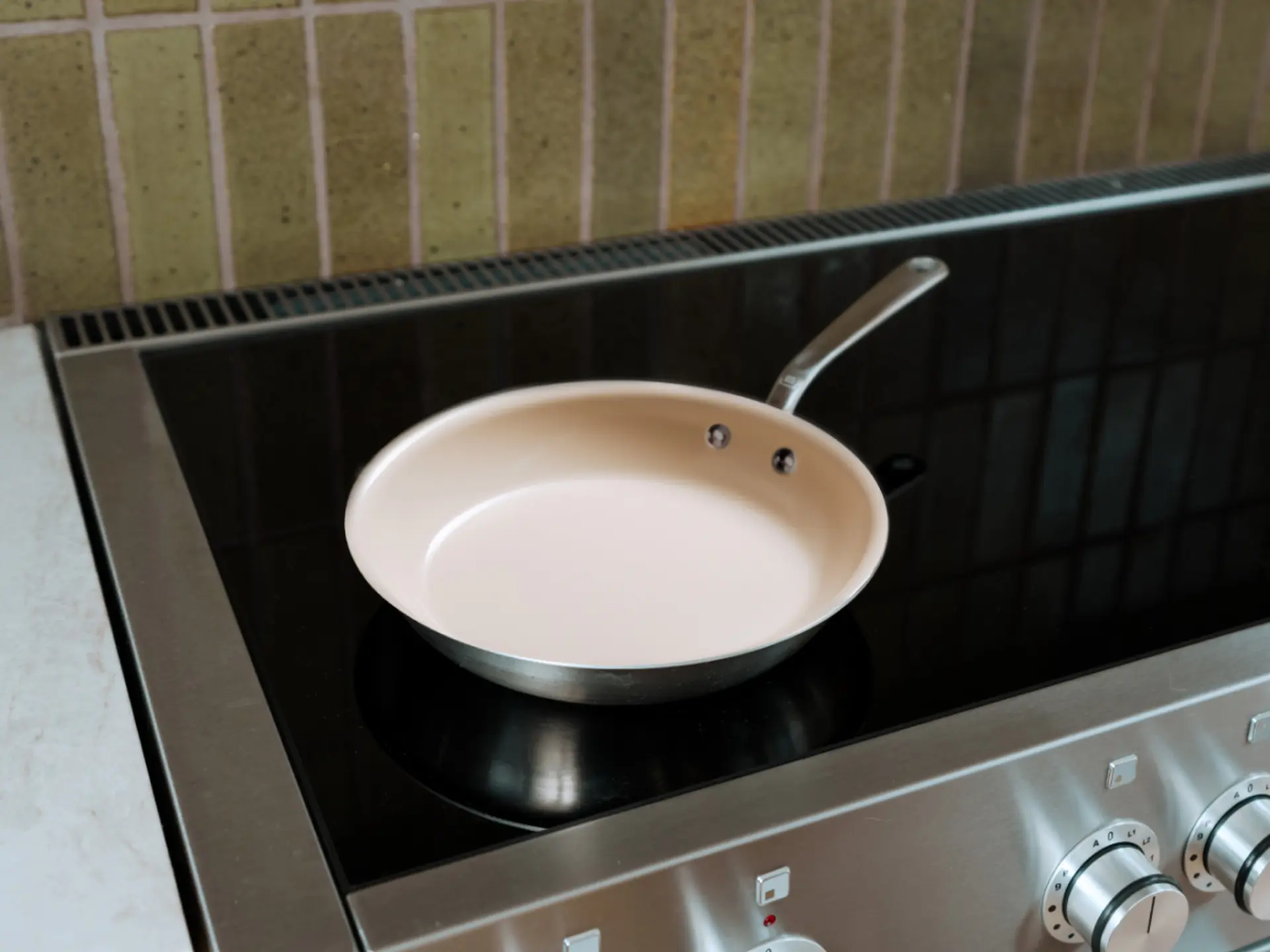 A pink frying pan with a silver handle sits on a black induction cooktop against a tiled backsplash.