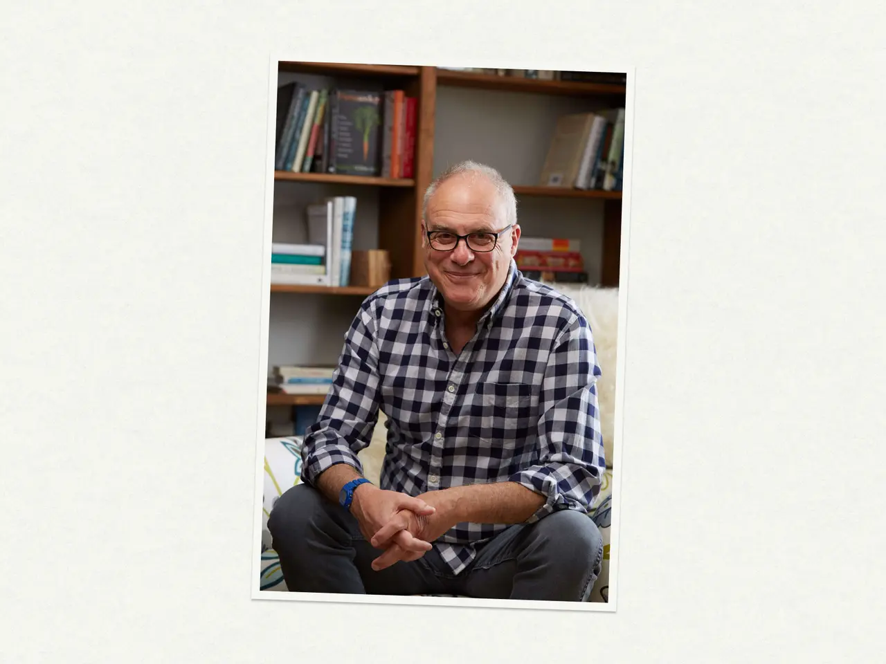 A smiling man wearing glasses and a checkered shirt sits casually on a couch in a cozy, book-filled room.