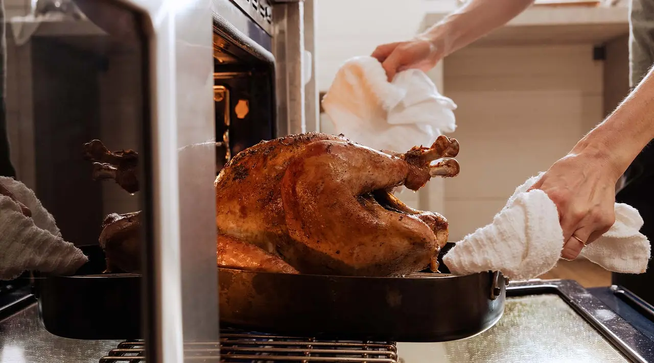 A roasted turkey being taken out of the oven by someone wearing oven mitts.