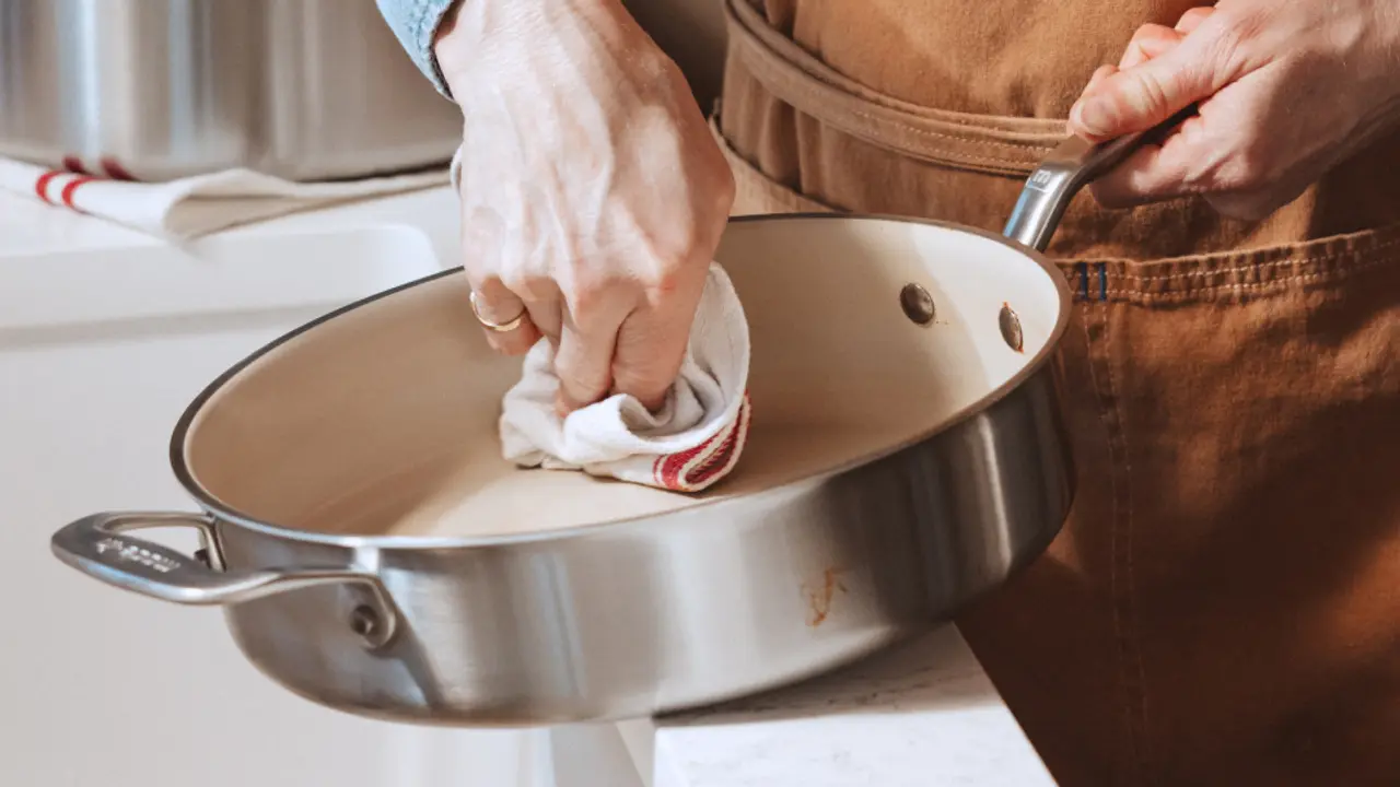 A person wearing an apron is cleaning a stainless steel cooking pan with a cloth.