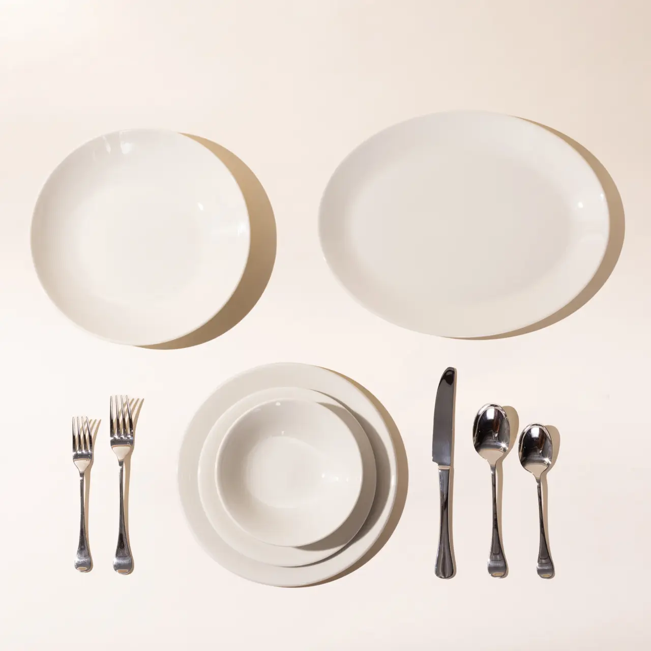 A neatly arranged dining set for two, with plates, bowls, forks, knives, and spoons on a plain background.