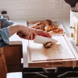 A person is using a knife to slice a piece of cooked chicken on a wooden cutting board in a kitchen.