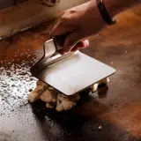 A hand holds a metal spatula while cooking cauliflower on a griddle.