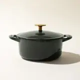 A dark-colored cooking pot with a lid featuring a contrasting gold-colored handle, set against a neutral background.