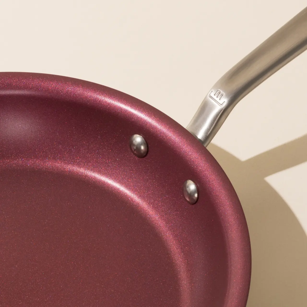A close-up of a maroon non-stick frying pan with a silver handle, highlighting the smooth surface and two rivets attaching the handle.