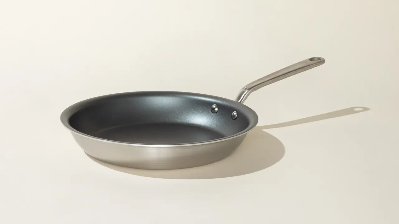 A stainless steel frying pan with a long handle is positioned on a neutral background.