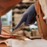 A person wearing blue gloves uses a "Made In" knife to fillet a fish on a cutting board.