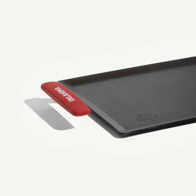 A black griddle with a red handle, designed for cooking, is shown at an angle with a spatula placed beside it.