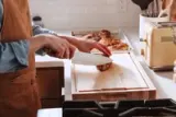 A person in an apron is cutting a piece of food on a wooden cutting board in a kitchen.