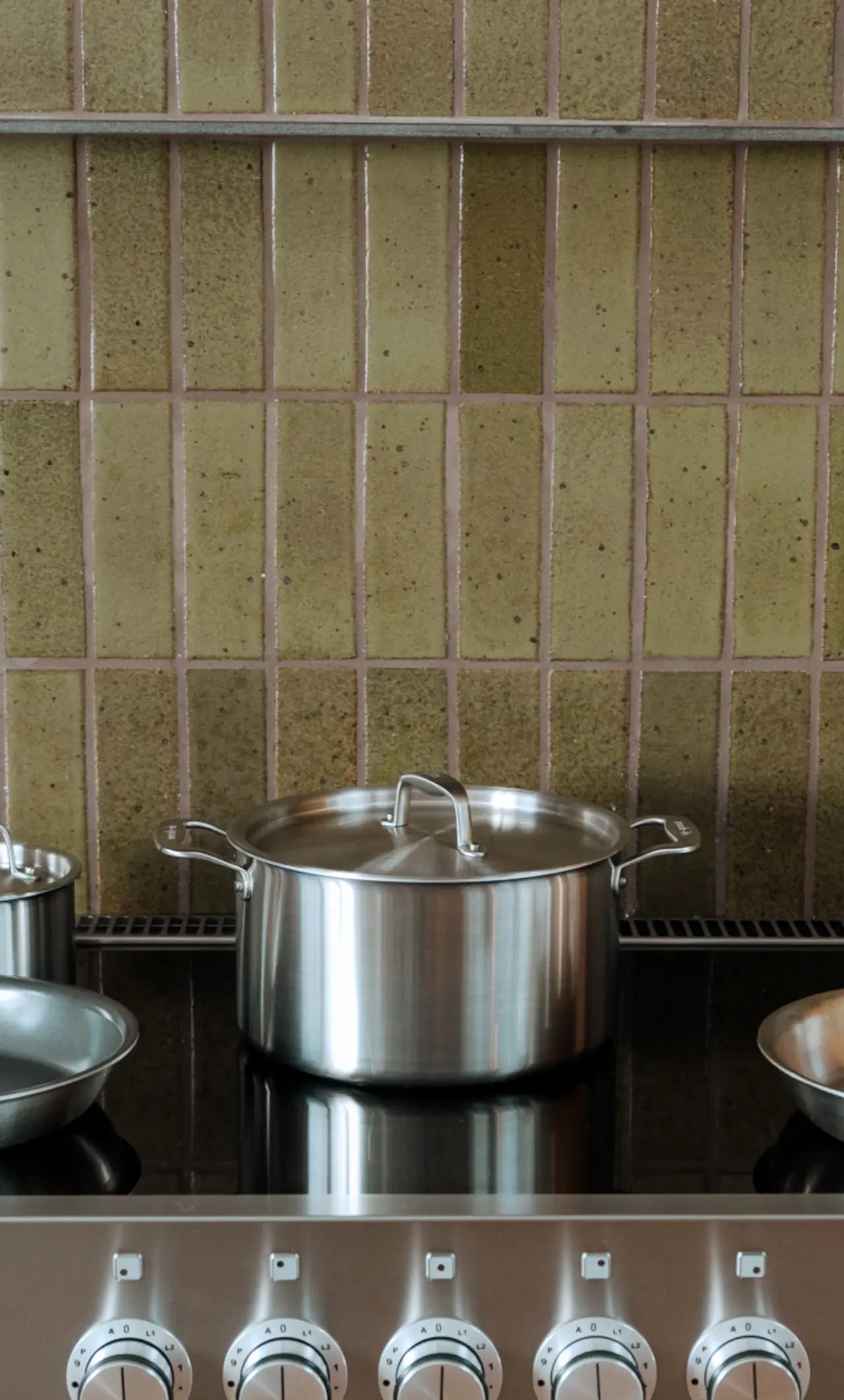 A stainless steel pot sits on a stove with control knobs in the foreground and a tiled wall behind.