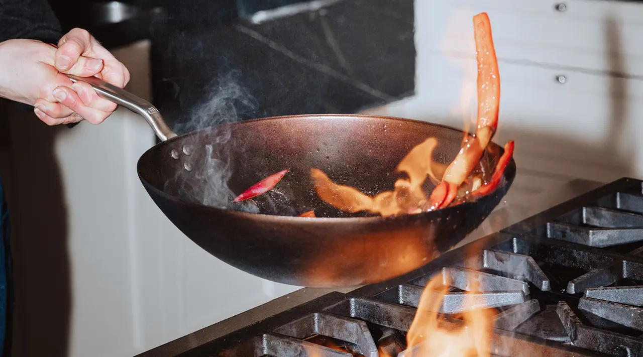 A person tosses colorful vegetables in a frying pan over an open flame in a kitchen setting.