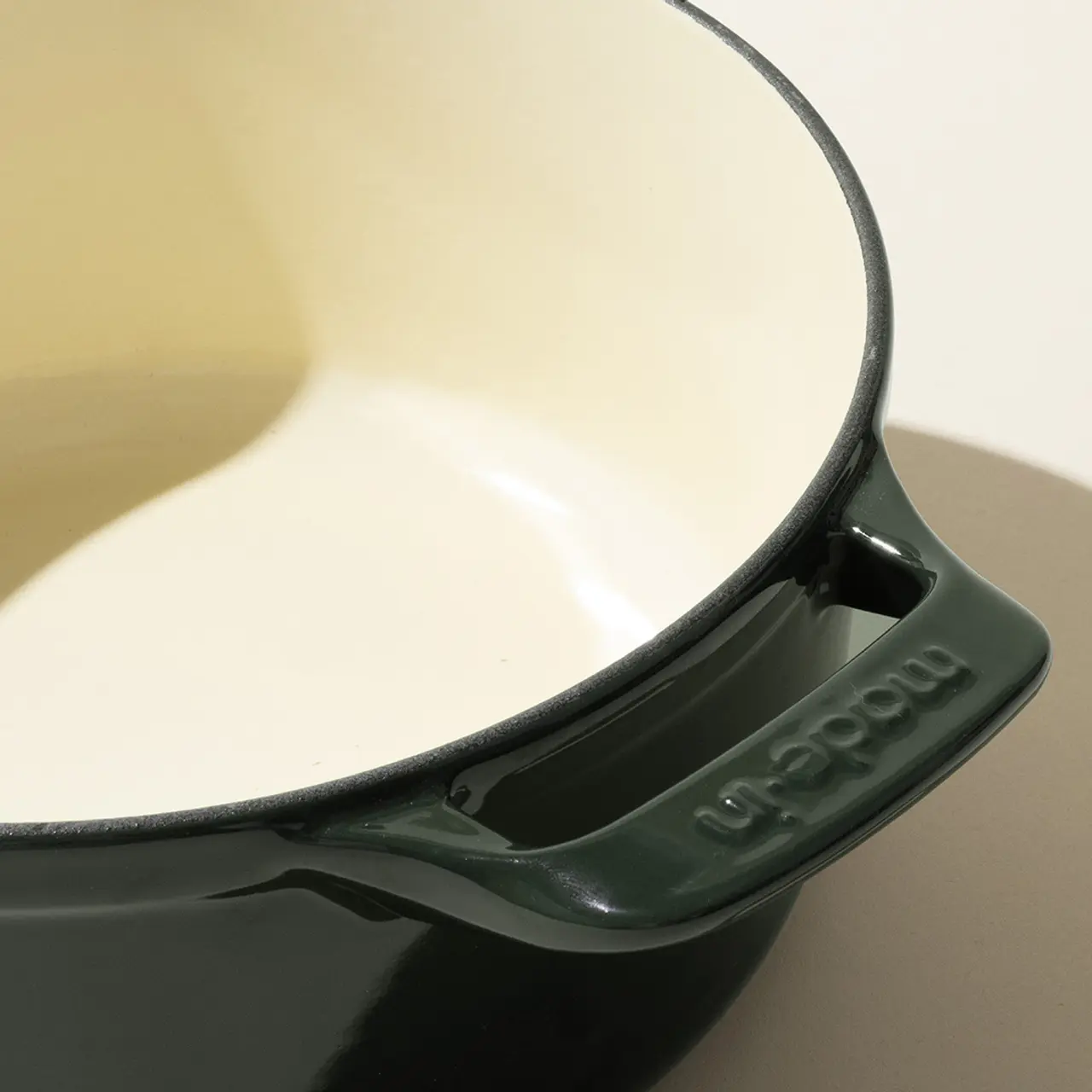 Close-up of a dark green enameled cast iron pot with a cream interior, highlighting the spout and brand imprint on the handle.