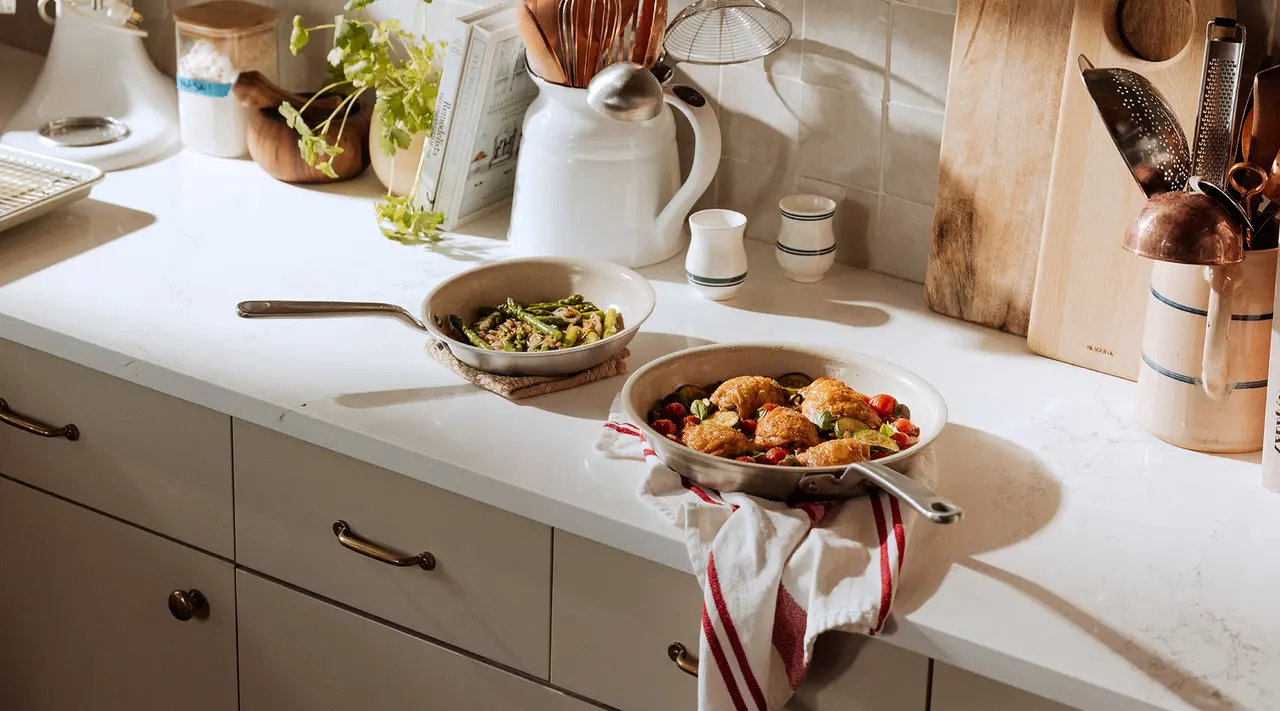 A neatly organized kitchen countertop displays cooking utensils and two pans of food, with a striped towel underneath one of the pans.