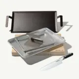 A collection of cookware and a knife neatly arranged on a surface, featuring a cast iron griddle, stainless steel pan with lid, a wooden cutting board, and a chef's knife.