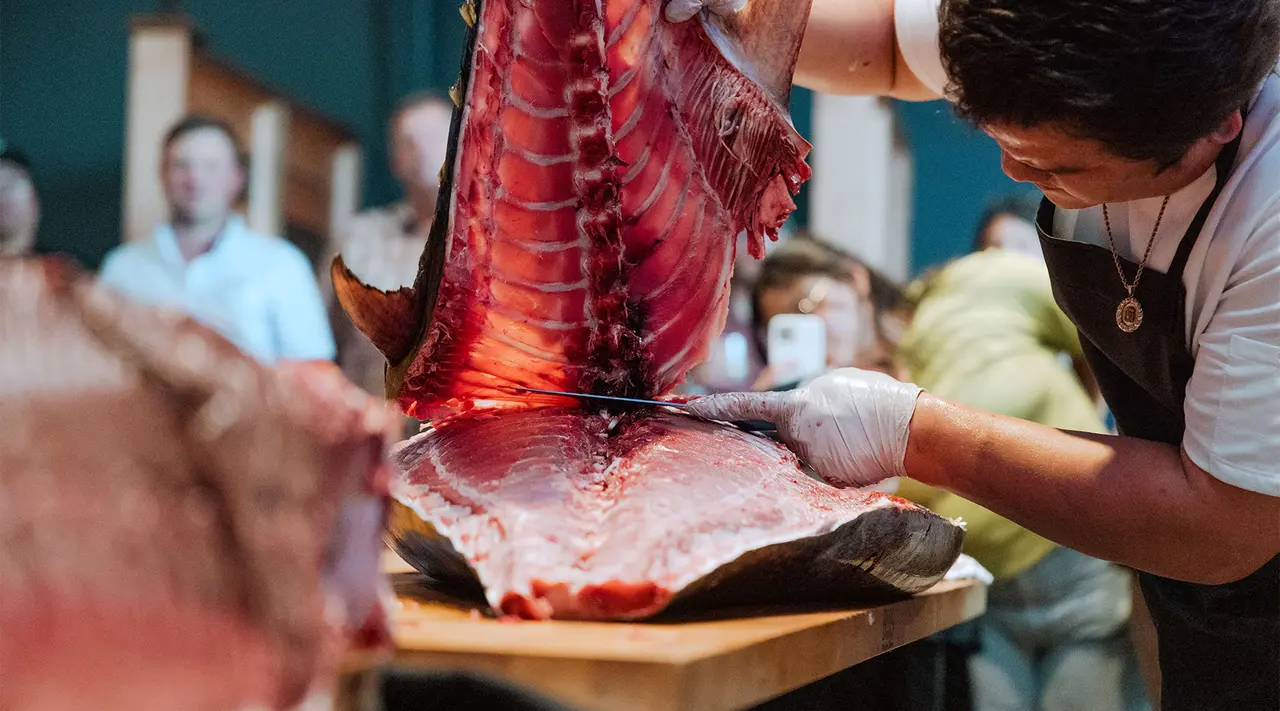 A person in a chef's apron is filleting a large fish on a table while onlookers watch in the background.