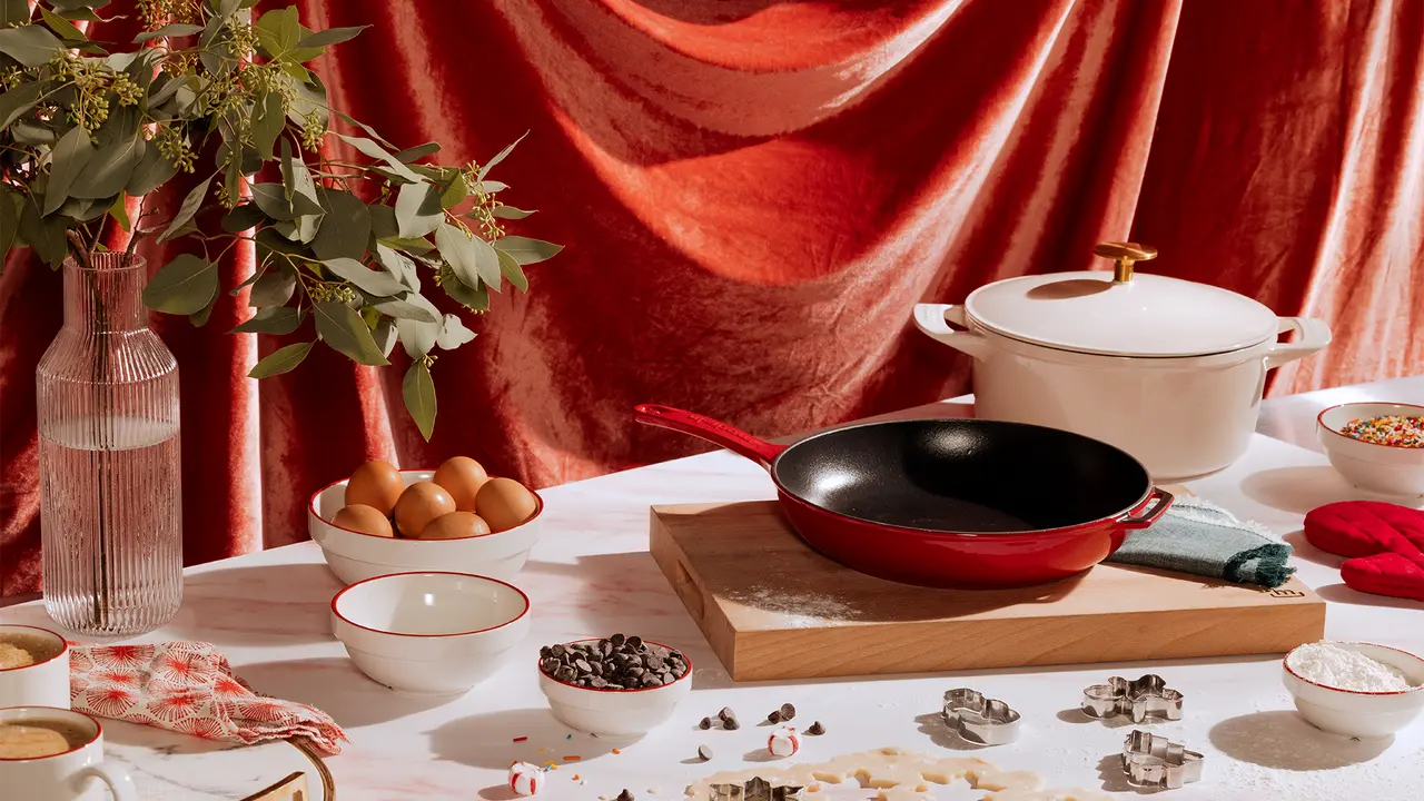 An elegantly set kitchen scene with cooking pots, a skillet, ingredients, and cutlery against a draped red backdrop.