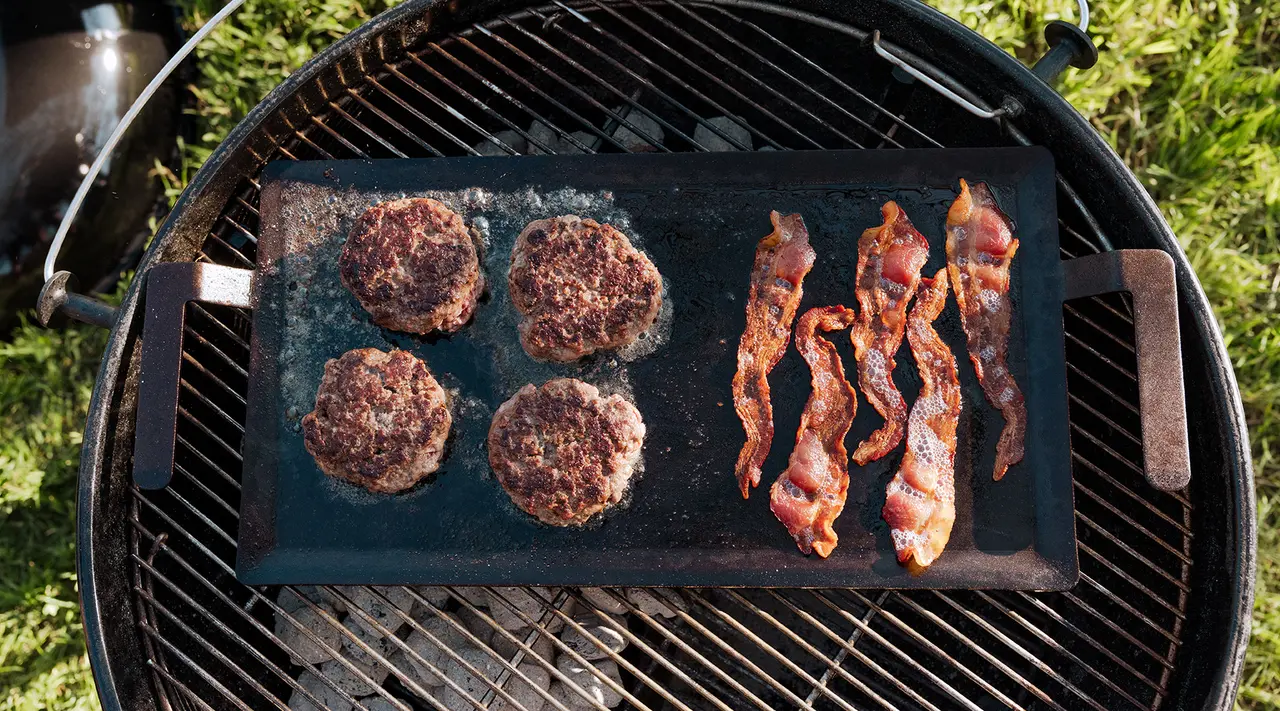 Hamburgers and bacon strips are sizzling on a flat griddle over a charcoal grill.