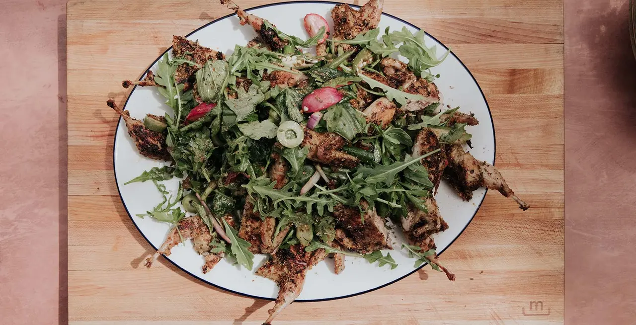 A plate of salad with mixed greens, some red slices that appear to be tomatoes, and pieces of grilled meat atop a wooden surface.