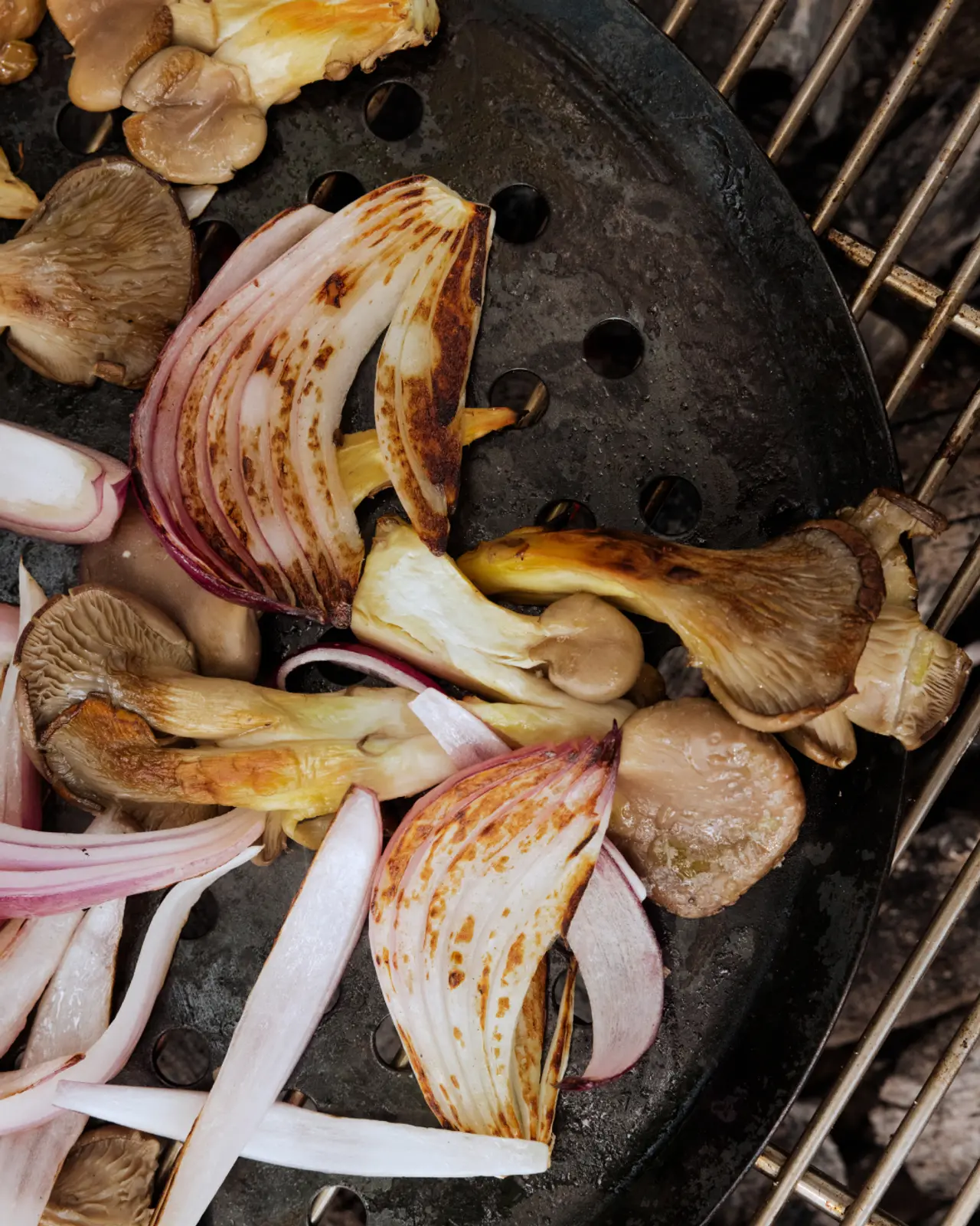 Sliced onions and mushrooms are grilled on a round, black perforated pan.