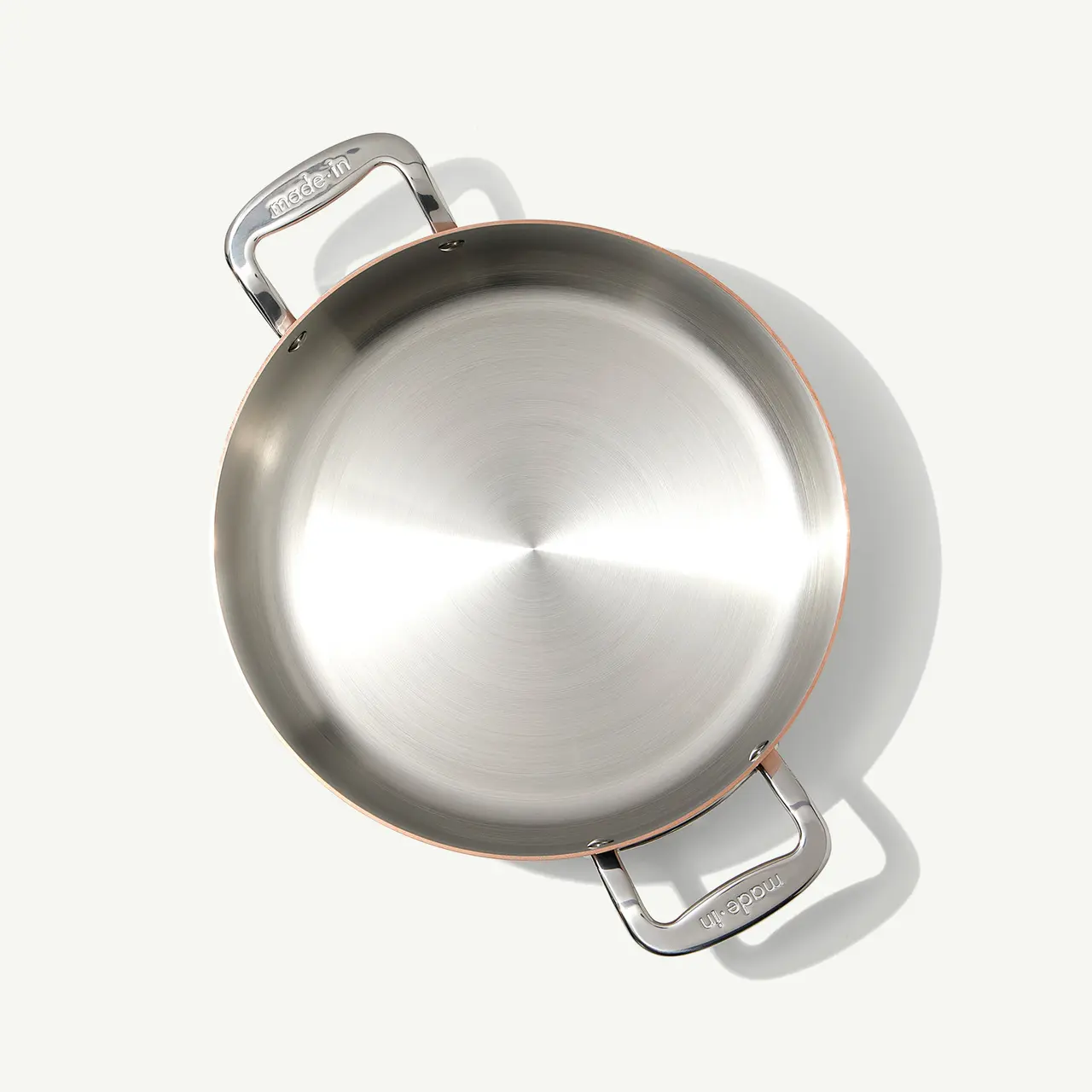 A stainless steel pan with a copper accent on the handle is viewed from above, casting a slight shadow on a light surface.