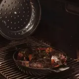 Sizzling steaks with herbs on an oval metal plate are being cooked on a barbecue grill.