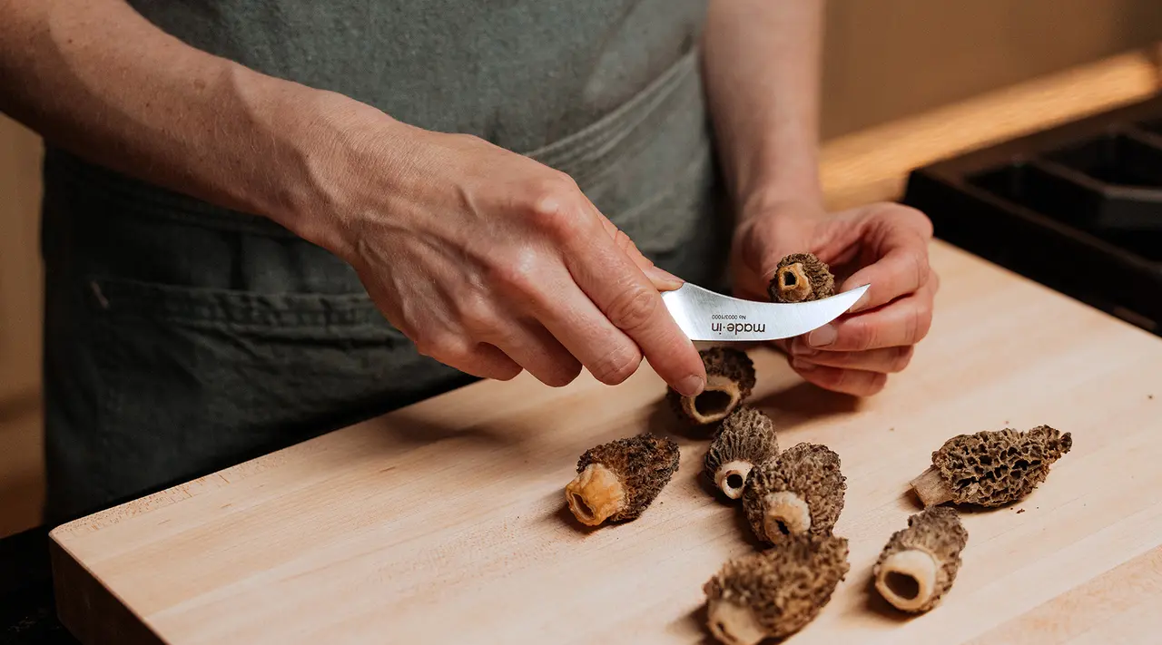 A person stands at a wooden countertop, preparing several morel mushrooms with a knife in hand.