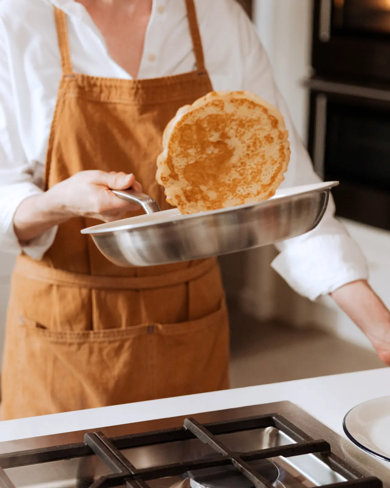 A person wearing an apron artfully flips a pancake in a kitchen setting.