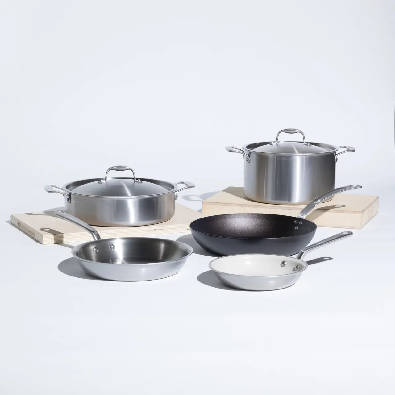 A collection of five cooking pans in various sizes and colors, including stainless steel and a black frying pan, arranged on a light background with wooden cutting boards.