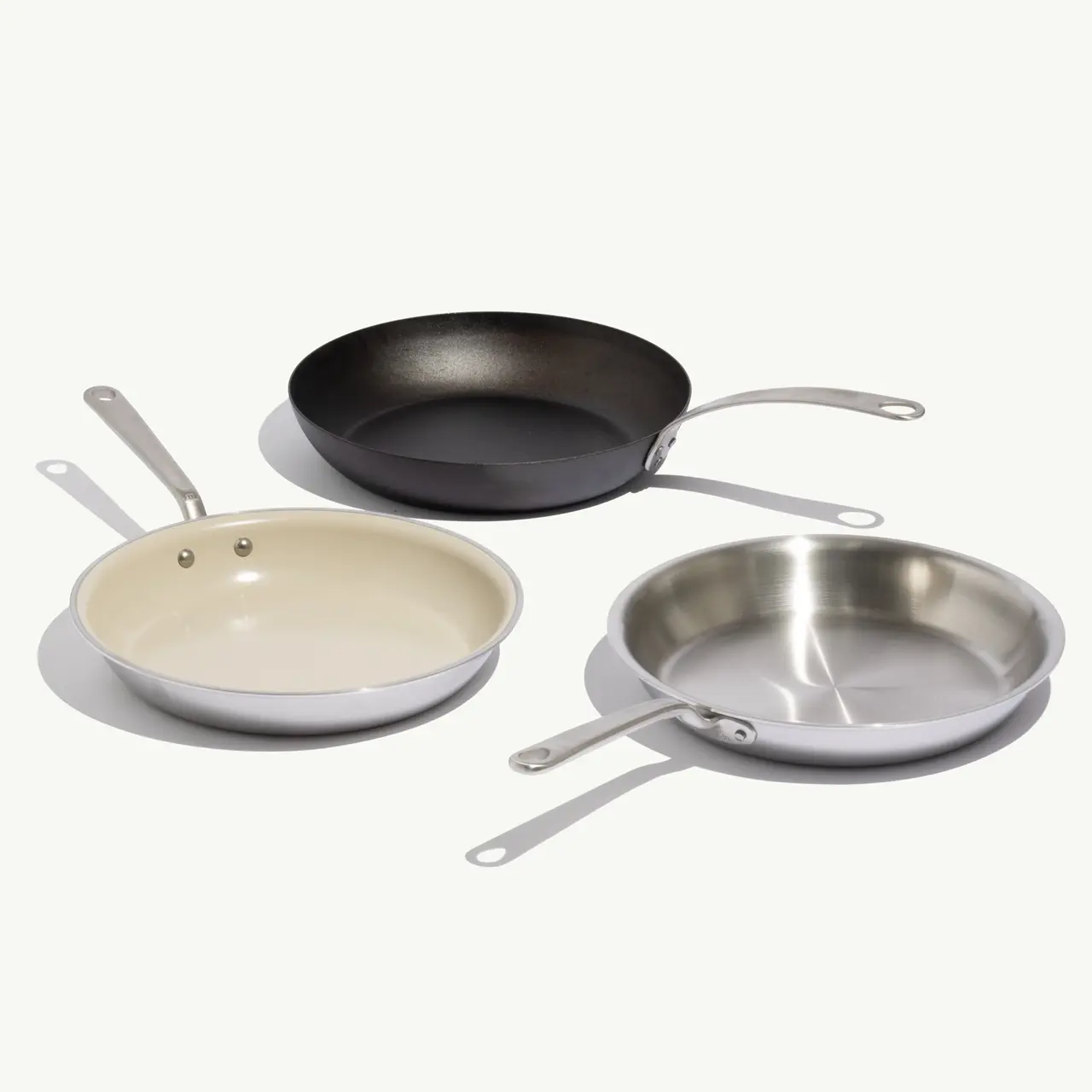 Three different frying pans in various materials—black cast iron, cream-coated, and stainless steel—arranged on a light background.