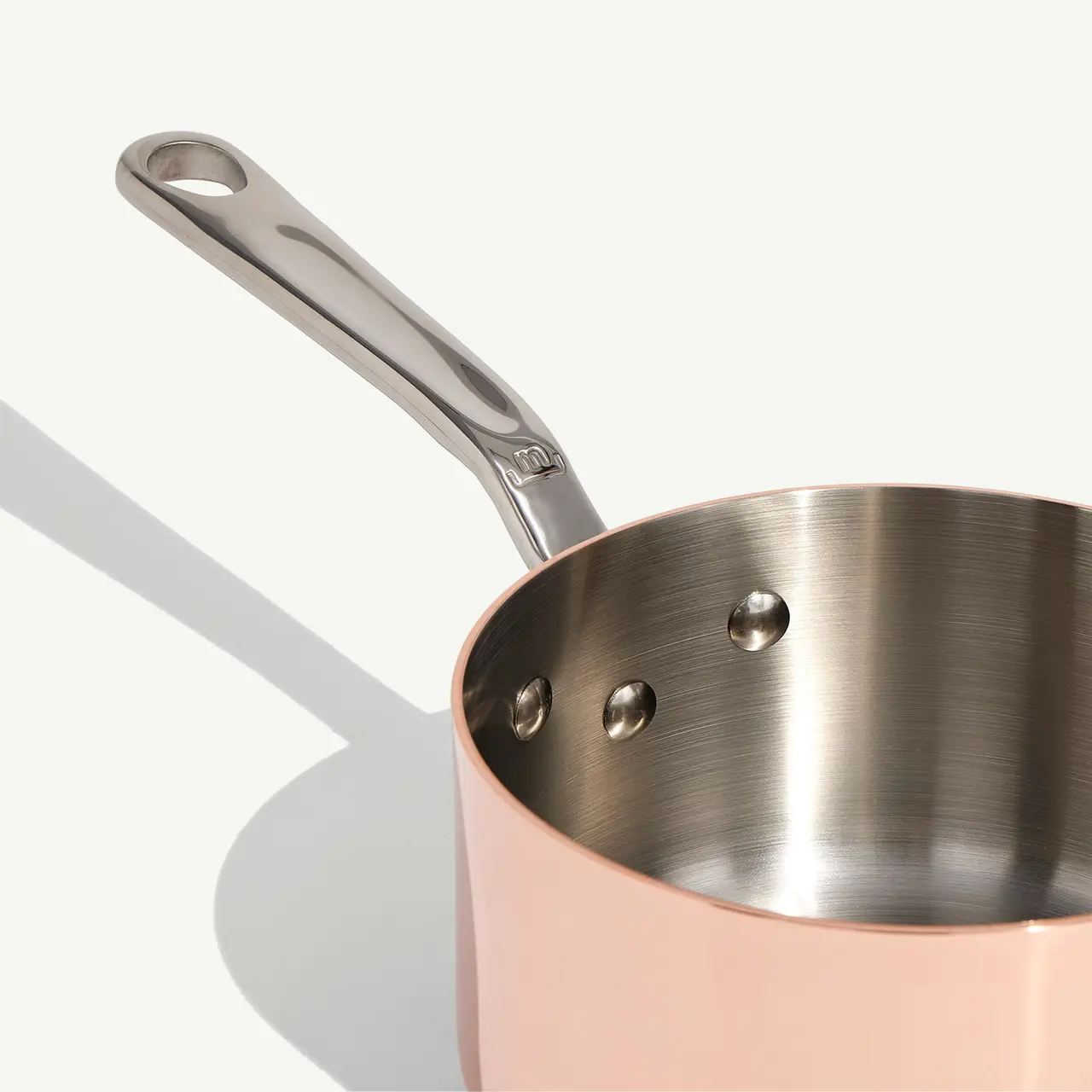 A stainless steel saucepan with a copper exterior and a long handle casts a shadow on a light background.