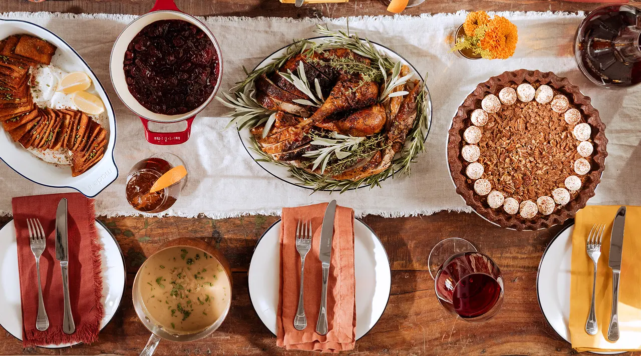 A festive dinner table from above, set with plates, cutlery, and a variety of dishes including a platter of grilled food, bowls with sides, a pie, and glasses of wine.