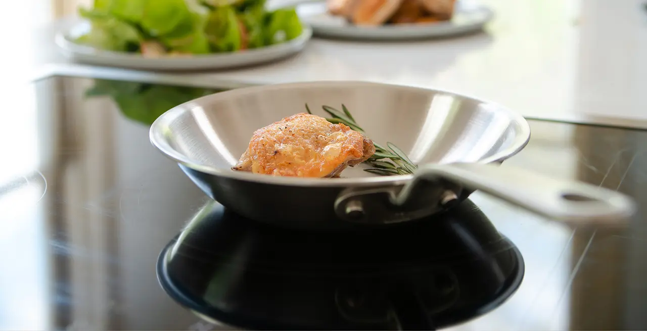 A piece of chicken with a sprig of rosemary is cooking in a stainless steel pan on a stovetop, with a salad in the background.
