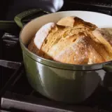 A freshly baked loaf of bread rests in a green cast iron Dutch oven on a stovetop.