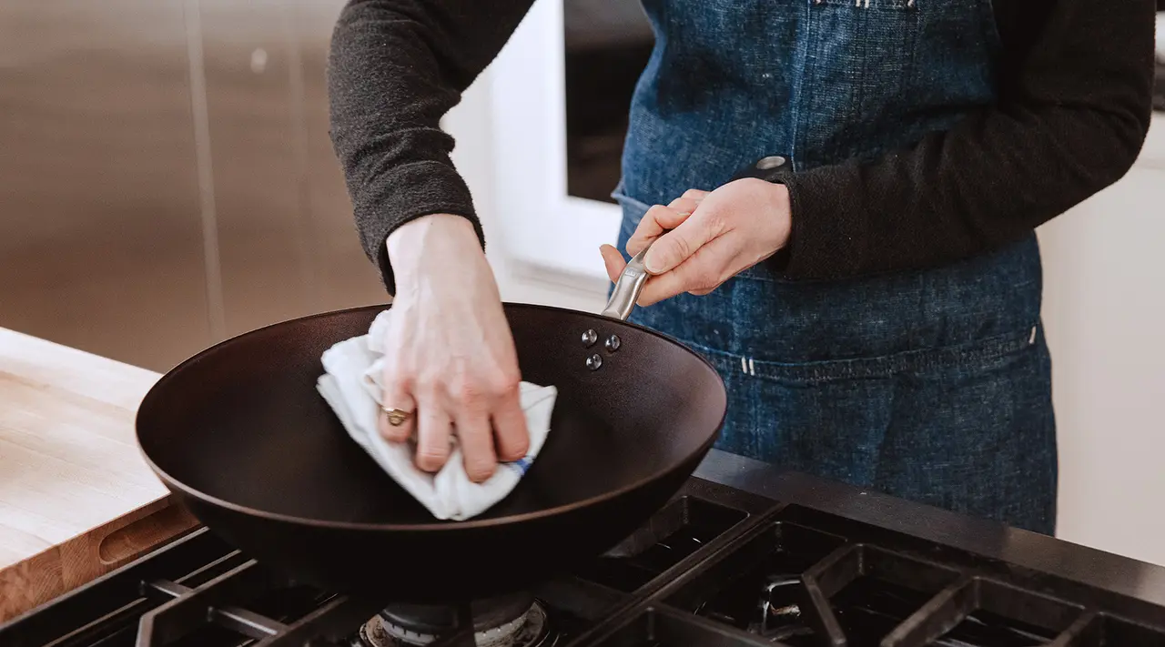 A person in a denim apron wipes down a black frying pan on a stove with a cloth.