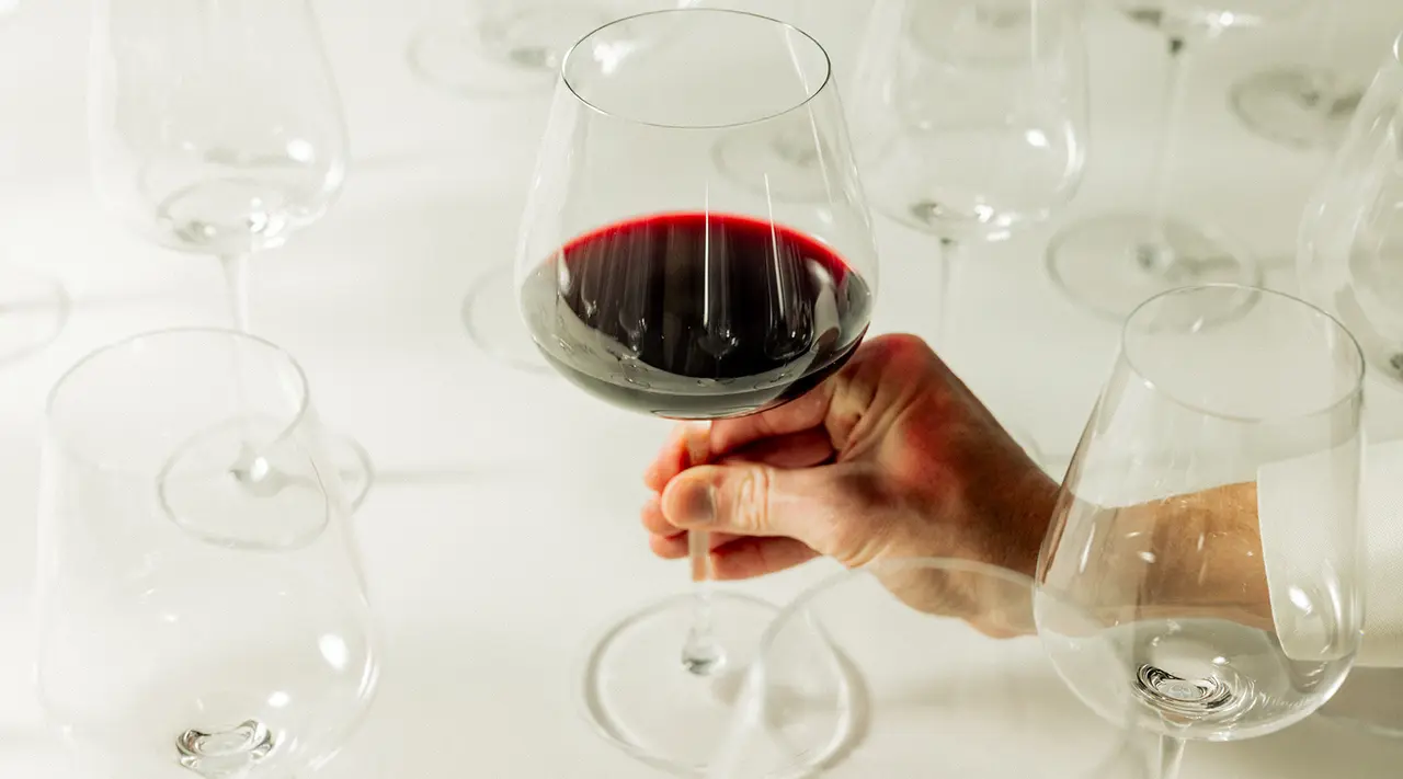 A hand holding a glass of red wine above a table scattered with empty stemware glasses.