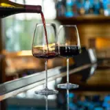 A bottle of red wine is being poured into a glass, with another filled glass beside it, against a bar setting with blurred background.