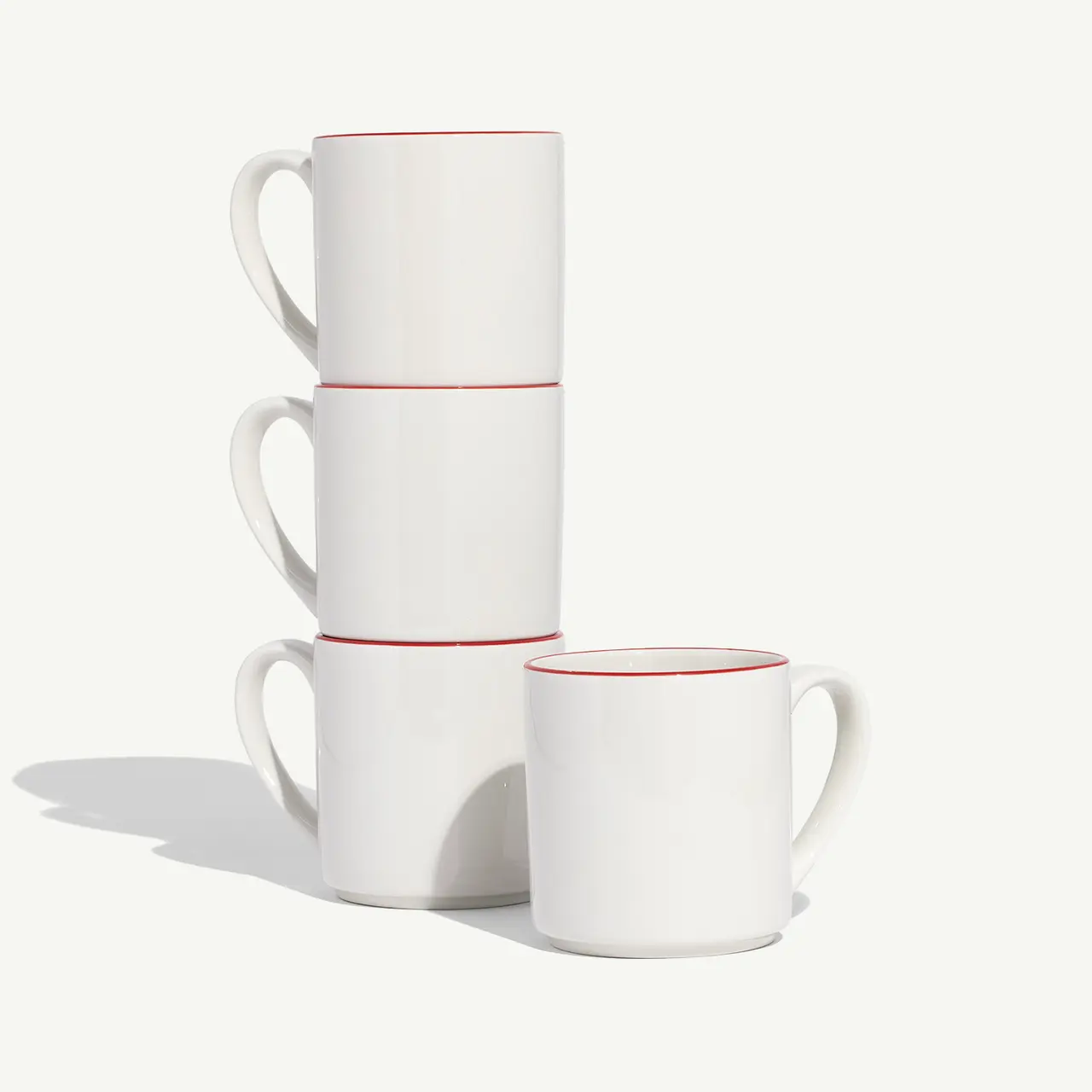 Three stacked white mugs with red rims are positioned next to a fourth mug against a plain background.