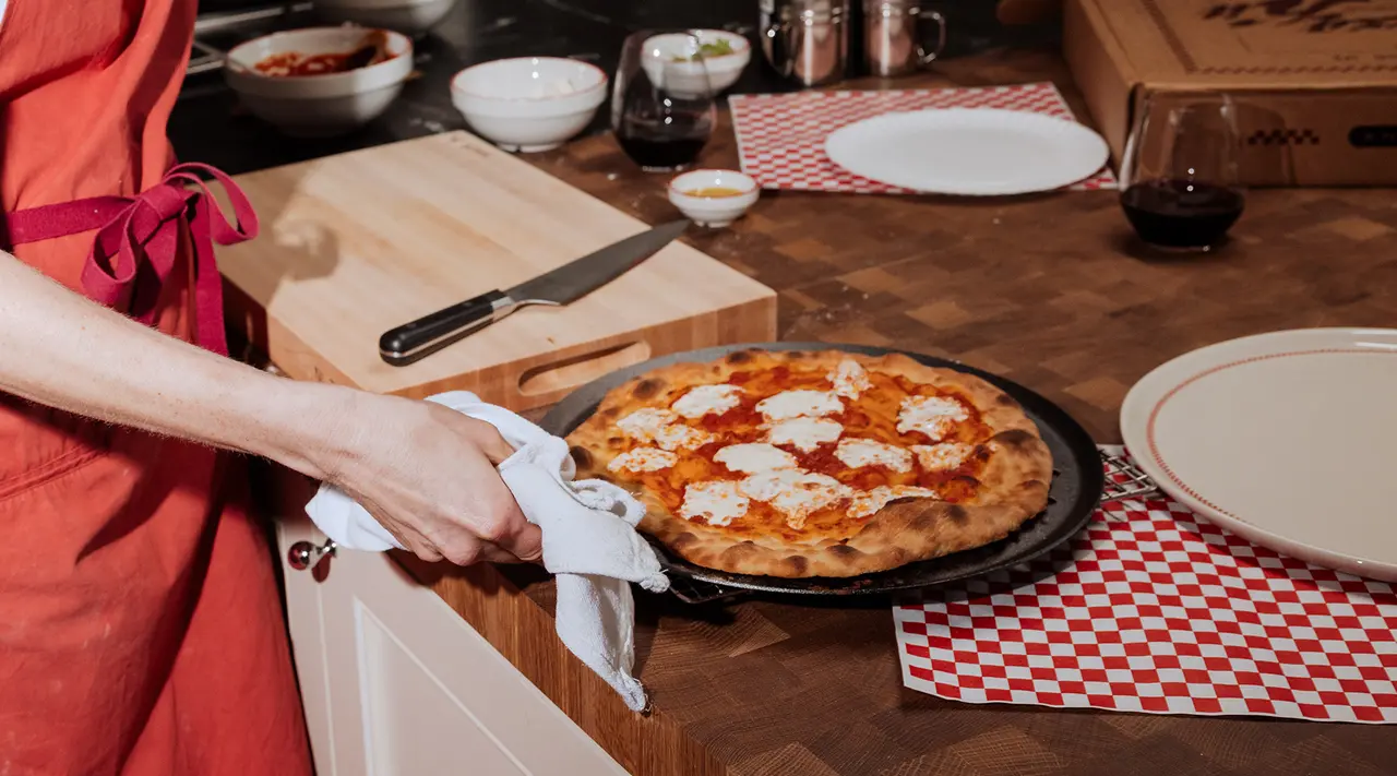 A person in an apron holds a freshly baked pizza on a tray while standing by a kitchen counter with various ingredients and cooking utensils around.