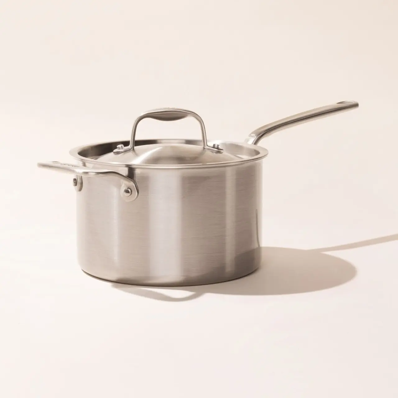 A stainless steel saucepan with a long handle is placed on a light-colored surface casting a soft shadow.