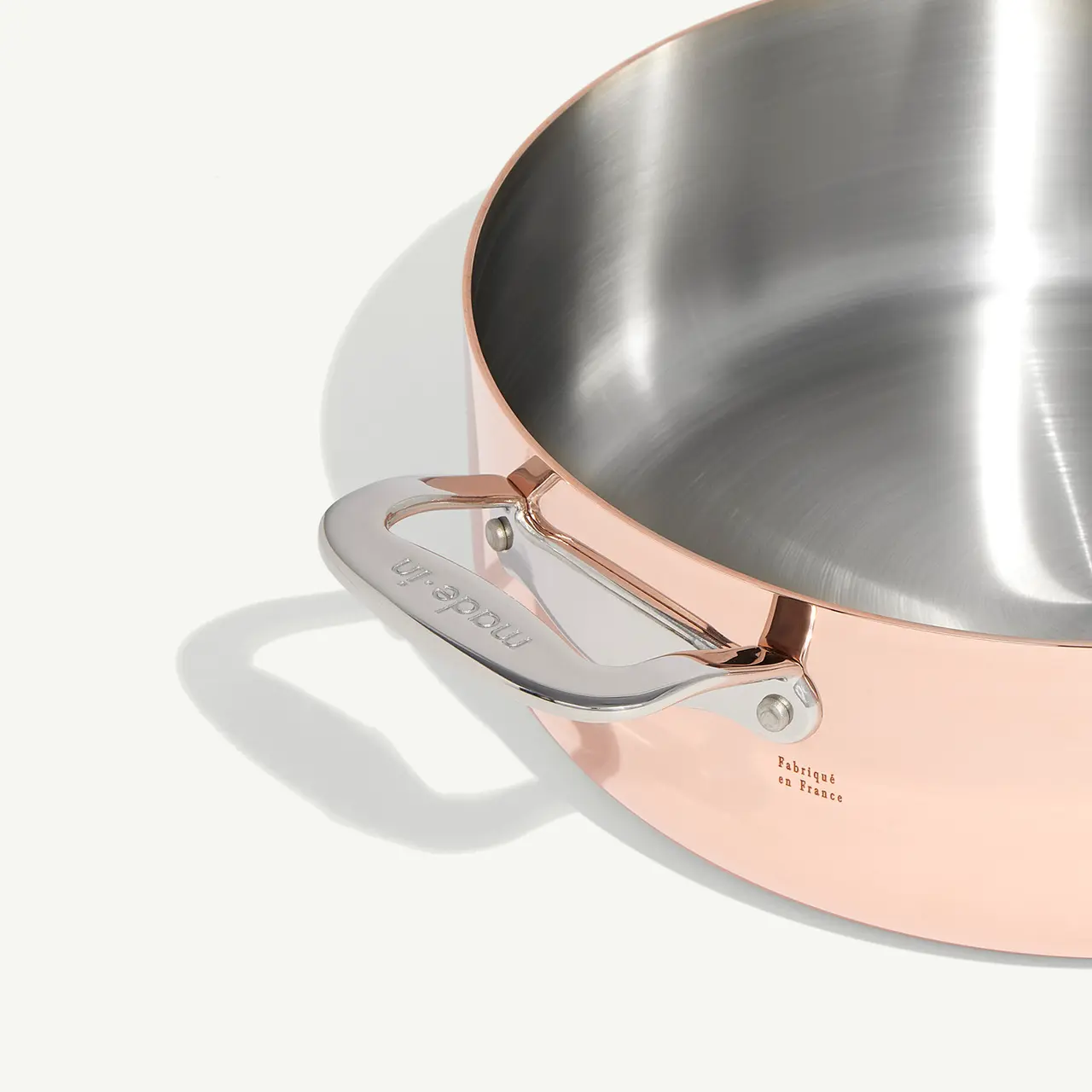 A stainless steel frying pan with a copper exterior and a detailed handle sits on a white surface, reflecting a soft light.