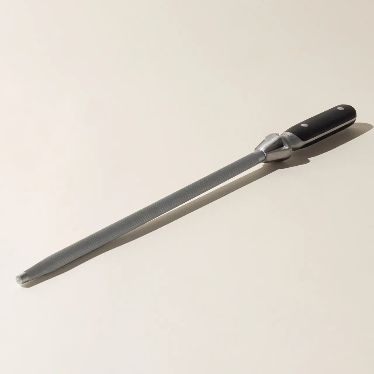 A long-handled sharpening steel lies on a plain surface, casting a soft shadow to one side.
